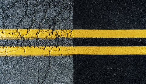 Directly above a newly paved asphalt road meeting an old worn-out crumbling road with freshly painted double yellow lines over both.